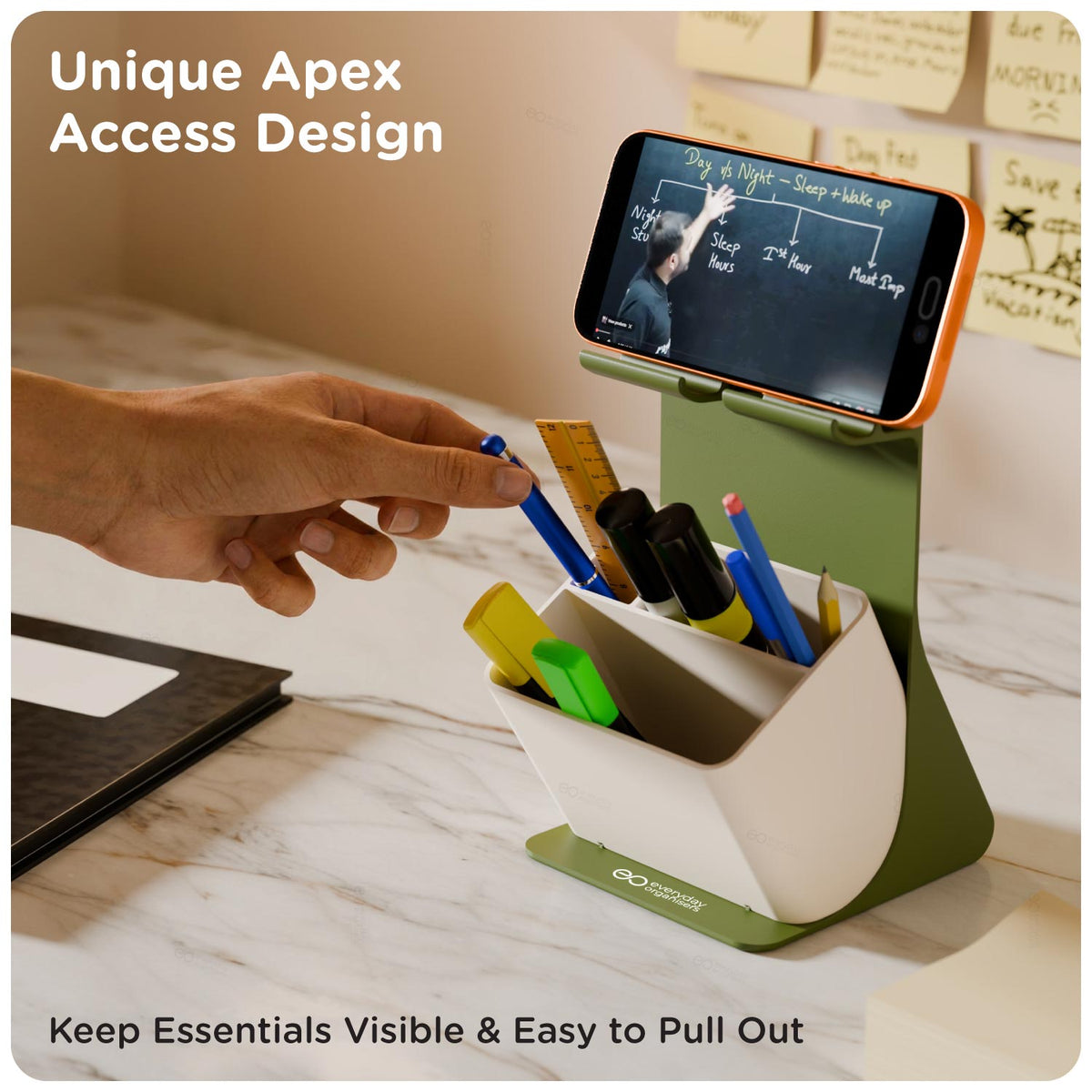 A green and white desktop organizer with a built-in phone stand, shown on a marble desk with a hand reaching toward pens in the cup. A smartphone is propped in the stand.